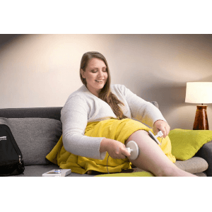 A smiling woman relaxing on sofa at home, applying Deep Oscillation therapy to her own lower leg using two handheld circular applicators.