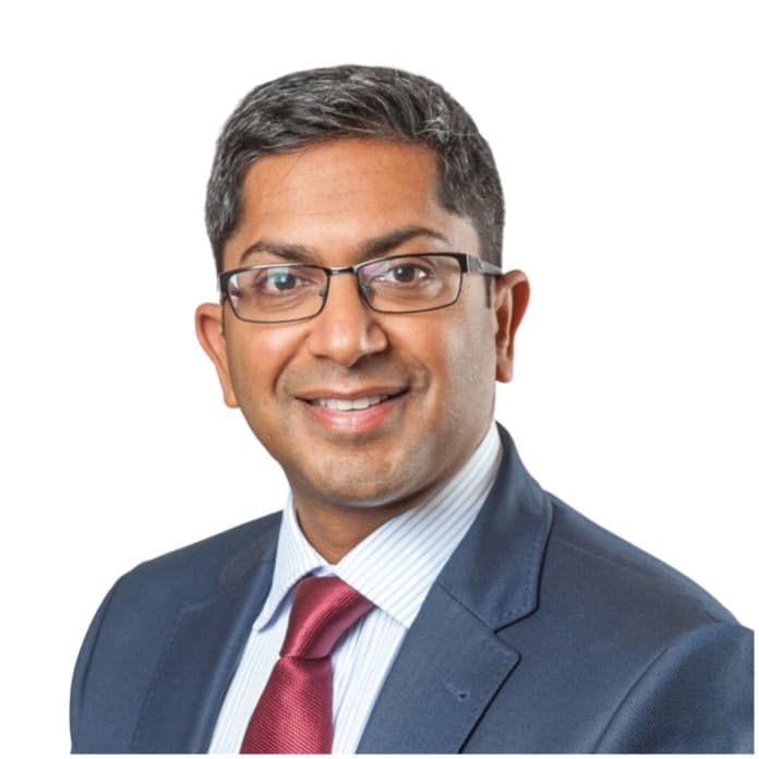A professional headshot of Mr Vasu Karri, who is smiling and wearing glasses, a blue suit, a light blue striped shirt, and a red tie.